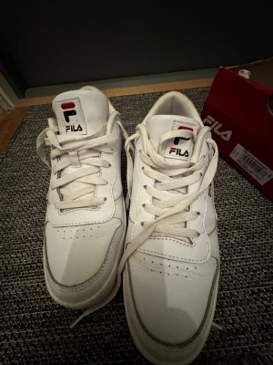 Vita Fila sneakers med logga - Klassiska vita sneakers från Fila med lågt skaft och tydlig Fila-logga på plös och sida. Skorna har vita snören, diskreta röda och blå detaljer samt en chunky sula. Tillverkade i syntetmaterial och läderimitation för en clean och sportig look.