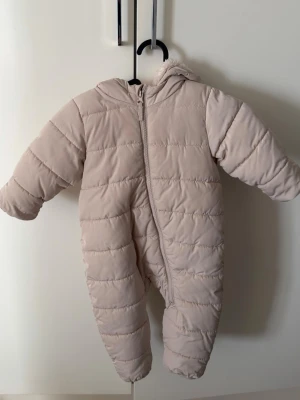 Beige overalljacka från H&M - Mysig beige overalljacka från H&M med huva och fluffigt foder. Jackan är quiltad och har dragkedja framtill som går snett ner mot benet. Perfekt för kalla dagar och håller dig varm och bekväm.