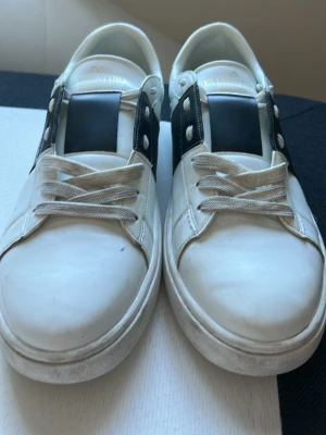 Valentino Garavani vita sneakers - Snygga vita sneakers från Valentino Garavani med en bred svart detalj över sidan. Skorna har klassisk låg profil och är tillverkade i läder med coola nitar på hälen. Perfekt för dig som vill ha en stilren och exklusiv look. Pris kan diskuteras 