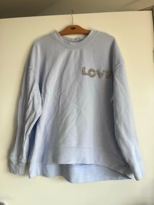 Ljusblå sweatshirt med glittertext - Ljusblå sweatshirt med rund hals och lång ärm. På bröstet står det 'LOVE' i stora bokstäver med glitterdetaljer. Tröjan är gjord i mjukt bomullsmaterial och har en något längre bakdel för en avslappnad look. Har små fläckar på magen.