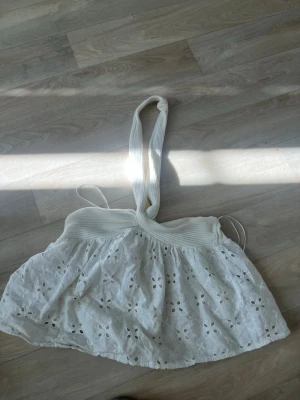 Vit topp med broderade detaljer - Söt vit topp l med bred ribbad resår i midjan och broderade blommönster över hela kjolen. Kjolen har en luftig och lätt form, perfekt för varma dagar. Den är gjord i bomull och har en romantisk känsla med fina spetsdetaljer.