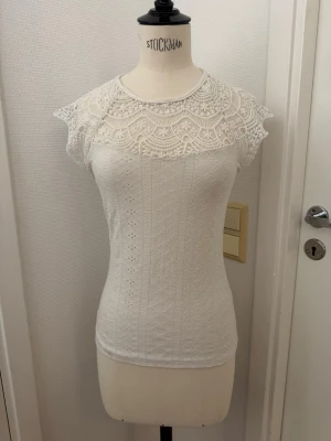 Vit topp i spets med rund hals - Supersöt vit topp med broderad spets upptill och små hålmönster över hela tyget. Toppen har korta ärmar, rund halsringning och knapp i nacken. XS stretch i tyget. Skickas eller hämtas i Hägersten 🤍