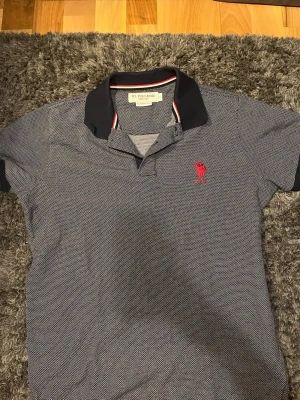 Mörkblå piké från U.S. Polo Assn - Snygg mörkblå pikétröja från U.S. Polo Assn med diskret mönster och röd logga på bröstet. Klassisk krage med vit och röd detalj på insidan. Perfekt till jeans eller shorts för en avslappnad look.
