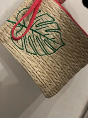 Beige kuvertväska med grön bladbrodyr - Säljer en beige kuvertväska i flätad look med grön broderad monstera-blad på framsidan. Väskan har rosa dragkedja och handledsrem, samt naturfärgad insida. Perfekt för att addera lite färg till din outfit!