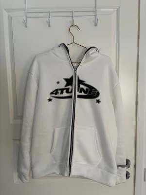 Vit hoodie med tryck från 4Tune - Säljer en vit hoodie från 4Tune med svart tryck och stjärndetaljer på bröstet samt coolt cirkelmönster med text på ryggen. Dragkedja framtill, två fickor och huva. Perfekt för dig som gillar streetwear och vill sticka ut lite extra.