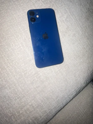 Blå iPhone 12 med dubbla kameror - Säljer en blå iPhone 12 med stilren design och rundade kanter. Baksidan är i glas och mobilen har två kameror samt Apples logga i mitten. Framsidan har en stor skärm som täcker nästan hela ytan. Perfekt för dig som gillar snygg teknik och vill ha en modern smartphone.