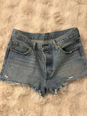 Levi's ljusblå jeansshorts med fransar - Säljer ett par klassiska ljusblå högmidjade jeansshorts från Levi's med råa fransade kanter och slitna detaljer. Shortsen har fem fickor, bälteshällor och Levi's ikoniska läderpatch bak. Perfekta för sommardagar och festivaler. Storlek W29. Använd fåtal gånger