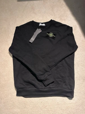 Svart sweatshirt från Stone Island - Säljer en svart sweatshirt från Stone Island med klassisk rund halsringning och ikonisk kompasspatch på bröstet. Tröjan har ribbade muddar vid ärmslut och nederkant samt en stilren look. Perfekt för dig som gillar streetwear och snygga detaljer.