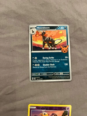 Pokémonkort Houndoom - Säljer ett Pokémonkort med Houndoom-motiv. Kortet har en färgstark illustration med eld och himmel i bakgrunden. Texten och detaljerna är tryckta på klassiskt glansigt pappmaterial. Perfekt för samlare eller Pokémonfans.