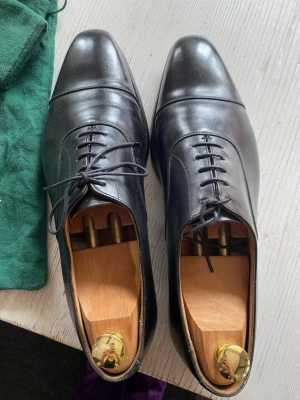 Svarta loafers från Crockett & Jones - Stilrena svarta loafers från Crockett & Jones i blankt läder. Klassisk design med rundad tå och diskreta sömmar över tån. Skorna har snörning och låg klack, perfekta för en dressad look. Levereras med träskoblock och grön tygpåse med logga.
