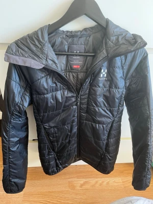 Svart pufferjacka från Haglöfs - Snygg svart pufferjacka från Haglöfs med huva och dragkedja framtill. Jackan har en lätt glansig yta och diskret logga på bröstet. Perfekt för kalla dagar och tillverkad i syntetmaterial som håller värmen. Stilren och enkel design som passar till det mesta.