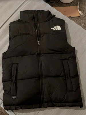 Svart dunväst från The North Face storlek S - Säljer en svart dunväst från The North Face med hög krage och dragkedja framtill. Västen har två fickor med dragkedja på utsidan och en innerficka. Klassisk logga på bröstet och 700-brodyr vid nederkanten. Perfekt för lager-på-lager och streetwear.