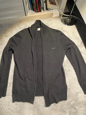 Mörkgrå ziptröja från Gant - Mörkgrå ziptröja från Gant med hög krage och diskret logga på bröstet. Tröjan har långa ärmar, ribbade muddar och är tillverkad i mjukt stickat material. Perfekt för lager på lager och enkel att matcha med olika outfits. 8,7/10 skick