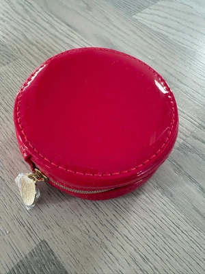 Rund röd plånbok/sminkväska med dragkedja - YSL - YSL -  Snygg rund plånbok i blankt rött material med guldfärgad dragkedja och dekorativ dragkedjetag. Perfekt för kort, mynt eller småsaker. Den kompakta formen gör den enkel att ta med i väskan. Oanvänd 