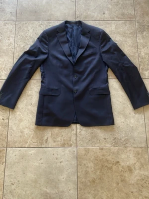 Mörkblå klassisk kavaj herr - Snygg mörkblå kavaj med två knappar framtill och klassiska slag. Kavajen har två fickor med lock och en innerficka. Fodrad insida och stilren passform, perfekt till skjorta och jeans eller kostymbyxor.