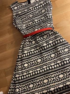Svartvit mönstrad klänning med rött bälte - Snygg svartvit klänning med grafiskt mönster och ett rött bälte i midjan. Ärmlös modell med rund halsringning och lätt utställd kjol. Perfekt för dig som gillar statement-plagg med retrovibbar.
