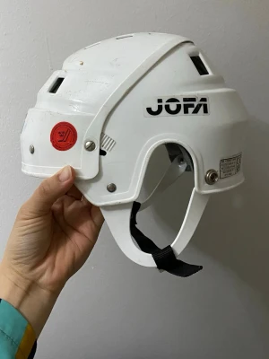 Vit hockeyhjälm från JOFA - Klassisk vit hockeyhjälm från JOFA med svart logga på sidan. Hjälmen har ventilationshål, justerbar hakrem och detaljer i metall. Tillverkad i tålig plast och godkänd enligt CE-standard. Passar perfekt för ishockey eller skridskoåkning.