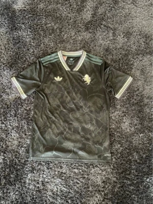 Grön Adidas t-shirt med blommönster - Snygg grön t-shirt från Adidas med diskret blommönster och guldfärgade detaljer på bröstet. Tröjan har korta ärmar, v-ringning och klassiska Adidas-ränder på axlarna. Perfekt för dig som vill ha en unik och sportig stil.