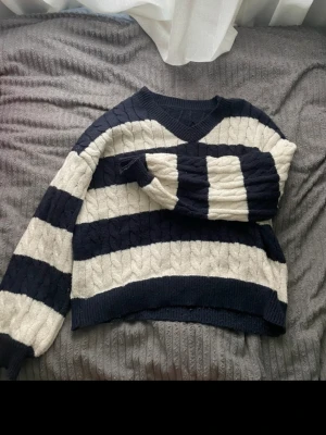 Randig stickad tröja i blått och vitt - Säljer en chunky stickad tröja med breda marinblå och vita ränder. Tröjan har V-ringning, lång ärm och ribbade muddar. Kabelstickat mönster ger extra struktur och en cool vintagekänsla. Perfekt för dig som gillar avslappnad och trendig stil.
