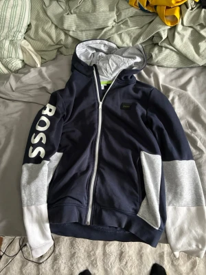 Mörkblå hoodie från BOSS med huva - Snygg mörkblå hoodie från BOSS med vit dragkedja och stora vita bokstäver på ena ärmen. Grå detaljer på ärmar och huva, samt en liten BOSS-logga på bröstet. Perfekt för dig som gillar stilrena och sportiga plagg.