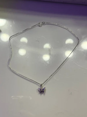 Silverhalsband med lila fjärilshänge - Supersött halsband i silverfärg med ett litet fjärilshänge i lila. Kedjan är tunn och smidig, perfekt för att ge en söt detalj till din outfit. Fjärilen har en glittrig känsla och ger ett lekfullt intryck.