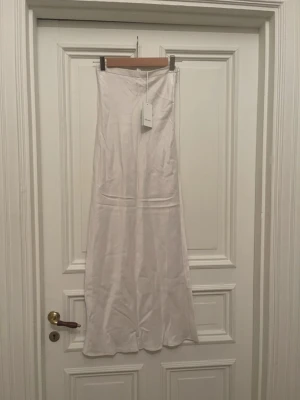 Vit satinkjol från Dagmar - Säljer en lång, vit satinkjol från Dagmar med slät midja och lätt utsvängd passform. Kjolen har en elegant glansig yta och dragkedja i sidan. Perfekt för dig som gillar stilrena och trendiga plagg.