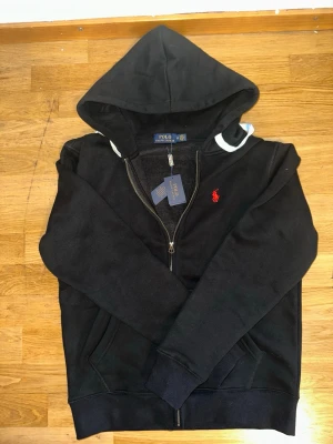 Svart hoodie från Polo Ralph Lauren - Svart hoodie med dragkedja från Polo Ralph Lauren. Klassisk design med huva, kängurufickor och rött broderat logotyp på bröstet. Mjuk bomullsblandning som är skön att bära. Perfekt för en avslappnad och stilren look.