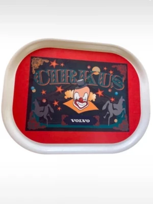 Cirkus-bricka Volvo med clownmotiv - Unik serveringsbricka med färgglatt cirkusmotiv, clown och hästar. Brickan är rektangulär med rundade hörn, vit kant och röd botten. Tillverkad i plast och har texten 'Cirkus' samt 'Volvo' på framsidan. Perfekt för dig som gillar retro och samlarprylar.