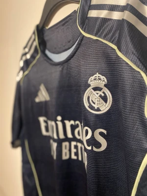 Real Madrid bortatröja 23/24 Bellingham 5 - Mörkblå Real Madrid bortatröja från Adidas säsongen 23/24. Tröjan har klubbmärke, Adidas-logga och sponsortryck i vitt samt ljusgula detaljer längs sidorna. Korta ärmar med tryck på båda. Storlek L och tryck med Bellingham nummer 5 på ryggen. UCL batch (denna är inne hos oss nu)
