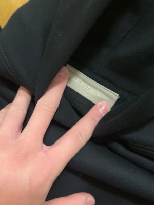 Svart Essentials hoodie med huva - Säljer en svart hoodie från Essentials med stor huva och magficka. Hoodien har diskret Essentials-logga på bröstet och är gjord i ett mjukt, tjockt material som känns riktigt skönt. Perfekt för dig som gillar en clean och stilren look.