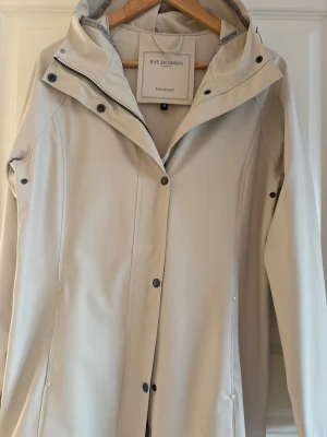 Beige regnjacka från Ilse Jacobsen - Snygg beige regnjacka från Ilse Jacobsen med huva och tryckknappar framtill. Jackan har en stilren design, vattentätt material och diskreta sömmar. Perfekt för regniga dagar när du vill hålla dig torr och se trendig ut.
