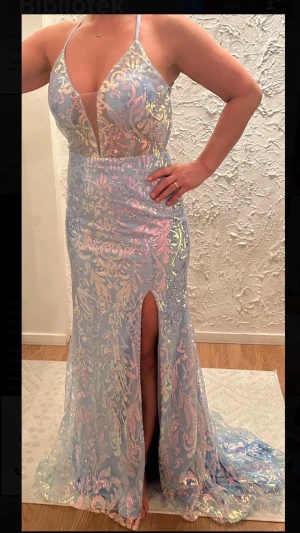 Ljusblå bal / fest klänning  -  glittrig långklänning i ljusblått med mönster. Klänningen har djup v-ringning, smala axelband, snörning i ryggen och en slits framtill. 