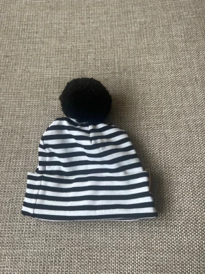 Randig mössa med svart tofs - Säljer en stilren mössa med svarta och vita horisontella ränder och en stor svart tofs på toppen. Mössan har en klassisk form och är tillverkad i mjukt tyg, perfekt för att hålla värmen och ge en trendig look.