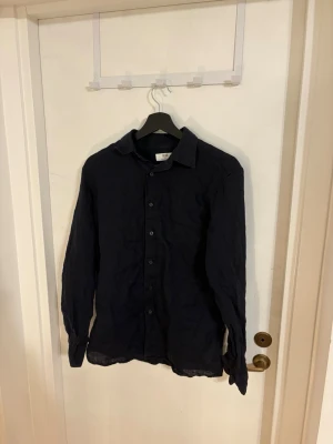Mörkblå linneskjorta från Uniqlo - Snygg mörkblå skjorta från Uniqlo i luftigt linne. Klassisk krage, långa ärmar och knappar hela vägen framtill. Perfekt för dig som gillar en enkel och stilren look.