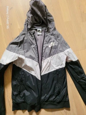 Nike vindjacka med huva, svart/grå - Nike vindjacka i svart, grått och vitt med dragkedja och huva. Jackan har två fickor med dragkedja och meshfoder på insidan. Snygg sportig design med Nike-logga på bröstet. Perfekt för blåsiga dagar.