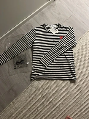 Randig tröja Comme des Garçons PLAY - Svartvit randig långärmad tröja från Comme des Garçons PLAY med det klassiska röda hjärtat på bröstet. Tillverkad i mjuk bomull och har rund hals. En ikonisk och stilren tröja som sticker ut med sin hjärtdetalj.