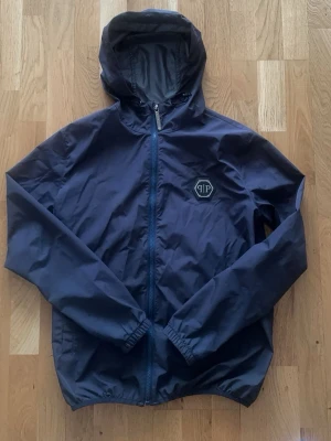 Mörkblå vindjacka från Philipp Plein - Skriv för kvitto bevis !!!!!Äkta!!!!  Kostar 9000kr helt ny Snygg mörkblå vindjacka från Philipp Plein med huva och dragkedja framtill. Jackan har elastiska muddar vid ärmslut och nederkant samt en diskret logga på bröstet. Tillverkad i lätt och vindtätt material, perfekt för blåsiga dagar.