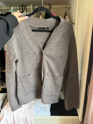 Beige kofta från 157 med fickor - Mysig beige kofta från 157 med två stora fickor framtill och en knappstängning. Koftan har ribbade muddar vid ärmslut och nederkant samt en djup V-ringning. Perfekt för lager på lager och enkel att matcha.