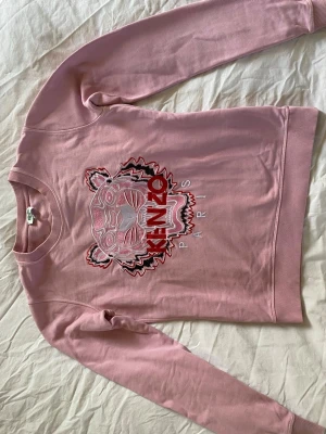 Rosa sweatshirt från Kenzo med tiger - Superfin ljusrosa sweatshirt från Kenzo med det ikoniska tigermotivet broderat framtill och texten 'KENZO PARIS'. Tröjan har rund hals och ribbade muddar. Svag fläck vid halsen och armhålan men annars i jättefint skick! 