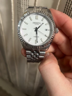 Silverfärgad klocka från Dissing - Stilren klocka från Dissing med vit urtavla, datumvisning och silverfärgad länk i metall. Klassisk rund form och tydliga markeringar på urtavlan. Perfekt accessoar för dig som gillar en clean och tidlös look.