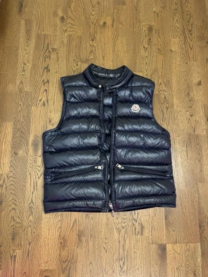 Mörkblå dunväst från Moncler -  Mörkblå dunväst från Moncler med glansig finish och hög krage. Västen har två dragkedjeförsedda fickor framtill och Moncler-logga på bröstet. Perfekt att bära över en t-shirt för en trendig look.