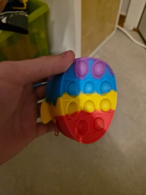 Färgglad pop it i silikon - Säljer en pop it-leksak i form av en figur, tillverkad i mjukt silikon. Den har färgglada ränder i lila, blått, gult och rött. Perfekt för att sysselsätta händerna och minska stress.