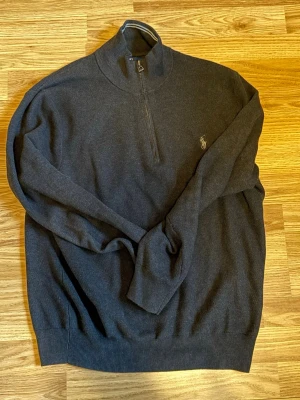 Mörkgrå stickad tröja Polo Ralph Lauren - Snygg mörkgrå stickad tröja från Polo Ralph Lauren med halv dragkedja. Tröjan har lång ärm, ribbade muddar och den klassiska broderade loggan på bröstet. Perfekt för dig som gillar stilrena och tidlösa plagg.