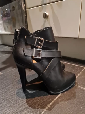 Svarta stövletter med spännen - Säljer ett par svarta stövletter med höga klackar och coola detaljer i form av dubbla spännen och remmar. Skorna har dragkedja bak och är tillverkade i ett läderliknande material. Perfekta för dig som gillar edgy och trendig stil.