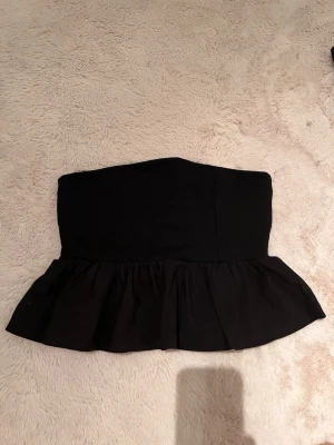 Svart axelbandslös volang tubtopp - Säljer en svart axelbandslös tubtopp med volangkant nedtill. Toppen har en croppad passform och är perfekt för dig som vill ha en stilren och trendig look. Tillverkad i mjukt tyg som sitter bekvämt mot kroppen.