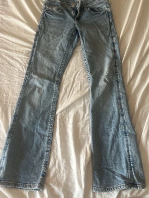 Ljusblå bootcut jeans med ficklock - Säljer ett par ljusblå bootcut jeans med klassisk femficksdesign och snygga ficklock bak. Jeansen har normalhög midja och är tillverkade i mjuk denim med lätt tvättad look. Perfekta för dig som gillar retrovibbar och avslappnad stil.