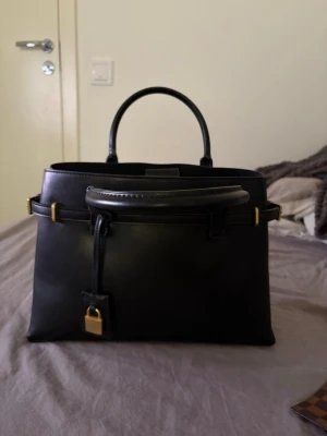 Svart handväska med gulddetaljer - Stilren svart handväska i slätt läder med guldfärgade metalldetaljer och lås. Väskan har ett topphandtag och en praktisk innerficka med dragkedja. Klassisk och elegant design som passar till många olika looks.