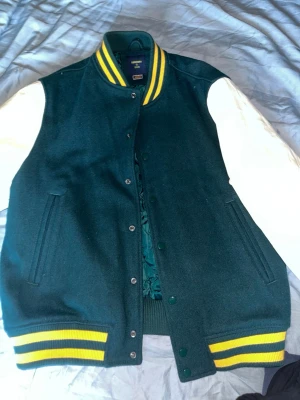 bomberjacka - Collegeinspirerad grön bomberjacka från Superdry med vita ärmar och gulrandiga detaljer på krage och muddar. Jackan har tryckknappar framtill, två fickor och quiltat foder. Perfekt för en sportig och avslappnad stil.