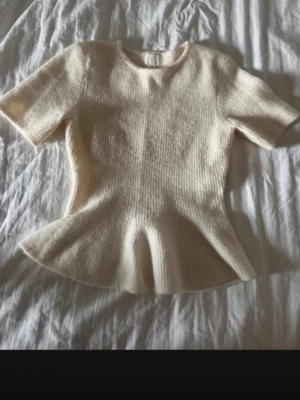 Vit ribbad topp från H&M - Supersöt vit ribbad topp från H&M med korta ärmar och peplumformad nederkant. Tillverkad i mjukt stickat material som ger en skön och snygg passform. Perfekt att styla med jeans eller kjol för en trendig look.