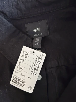 Svart skjorta från H&M Regular Fit - Svart skjorta från H&M med Regular Fit och klassisk krage. Skjortan har vita knappar framtill och på ärmsluten samt en bröstficka. Materialet är bomull och modellen är långärmad, perfekt för en clean och enkel look.